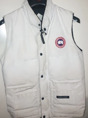 Vit dunväst från Canada Goose - Snygg vit dunväst från Canada Goose med klassisk logga på bröstet och två stora fickor framtill. Västen har hög krage, svarta tryckknappar och dragkedja. Perfekt för lager-på-lager och håller dig varm under kyliga dagar.