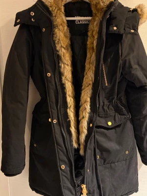 Svart parkas med fuskpäls - Svart parkas från Urbab Classic med fuskpäls längs huvan och insidan. Jackan har flera fickor med guldiga knappar och dragkedjor, justerbar midja och stor huva med pälskant som är avtagbat. Perfekt för kalla dagar. Rökfritt hem storlek L