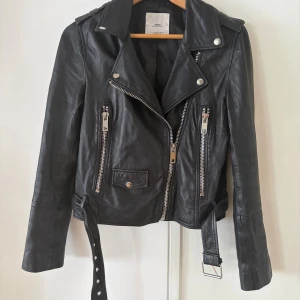 Svart äkta skinnjacka från Mango - SÅ snygg svart mjuk skinnjacka från Mango med klassisk bikerstil, silvriga dragkedjor, axelklaffar och bälte i midjan. Jackan har flera fickor med dragkedja och bred krage med tryckknappar. 