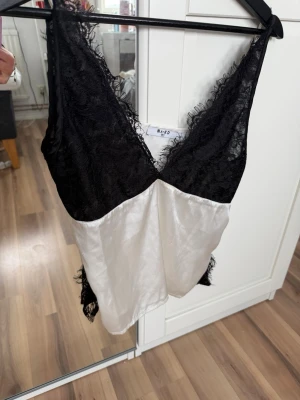 Svart och vit spetslinne från NA-KD - Snyggt linne från NA-KD med svart spets upptill och vit, glansig nederdel. Djup v-ringning och tunna, justerbara axelband. Perfekt för en chic och avslappnad look. Materialet känns som satin och spets, vilket ger en lyxig känsla.