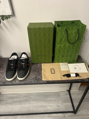 Gucci ace sneakers blå - Snygga svarta Gucci sneakers i GG Supreme canvas med klassiska röda och blå ränder på sidan. Skorna har vita sulor och svarta skosnören. Ikoniskt GG-mönster över hela skon och detaljer i läder. Perfekta för dig som vill ha en lyxig och trendig look.