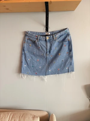 Blå jeanskjol Tommy Hilfiger storlek 30 - Cool ljusblå jeanskjol från Tommy Hilfiger med broderade bokstäver och små färgglada symboler. Storlek 30 ca 42-43 bred i midjan och 41cm i längden