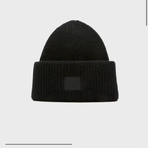 Svart ribbstickad mössa från Acne Studios - Säljer en svart ribbstickad mössa från Acne Studios med uppvikt kant och den klassiska Acne-loggan framtill. Mössan är gjord i mjukt material och har en enkel, stilren design som passar perfekt till kalla dagar.