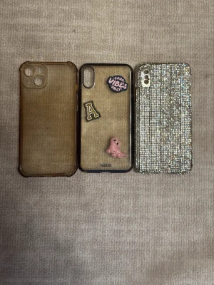Tre unika mobilskal i olika stilar - Säljer tre mobilskal: ett transparent brunt skal, ett genomskinligt skal från Holdit med dekorationer som 'Good Vibes Only', bokstaven A och en rosa figur, samt ett skal täckt av silvriga strass-stenar. Alla skal har öppningar för kamera och knappar.