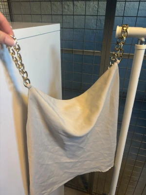 Beige linne med kedjerem - Trendig beige axelväska med bred guldfärgad kedjerem. har en mjuk, slouchy form och är tillverkad i ett tygmaterial som ger en avslappnad men ändå stylish look. Perfekt för att lyfta vilken outfit som helst.