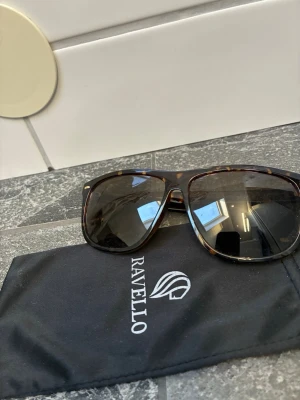 Bruna Ravello solglasögon - Snygga solglasögon från Ravello med klassisk brunmelerad båge och stora, fyrkantiga glas. Kommer med svart fodral med Ravello-logga. Perfekta för soliga dagar och ger en cool look. Liknar rayband boyfriend i stilen och formen!