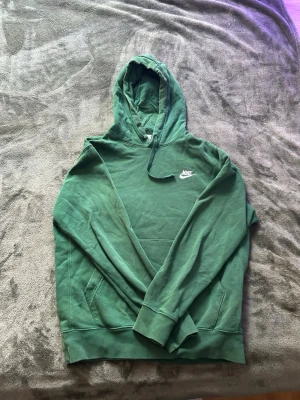Grön hoodie från Nike med huva - Snygg grön hoodie från Nike med klassisk logga broderad i vitt på bröstet. Hoodien har en stor ficka framtill, justerbar dragsko i huvan och ribbade muddar vid ärmar och nederkant. Tillverkad i mjuk bomullsblandning för extra komfort.