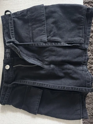 Svarta cargo kjol  - Säljer en svart cargo kjol  med stora fickor på sidorna och ett matchande tygbälte. Shortsen har dragkedja och knapp framtill och är perfekta för en avslappnad stil. Använd vid ett tillfälle 