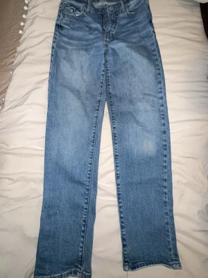 Blå Mom fit straight jeans  - Säljer ett par blå jeans med rak passform och klassisk femficksdesign. Jeansen har en ljus tvätt med subtila slitningar och är tillverkade i denim. Perfekta för dig som gillar en enkel och tidlös stil. Lite slitage vid ena knät