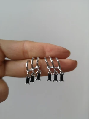 Silvriga örhängen med svarta stenar - Säljer ett par stilrena silvriga örhängen med rektangulära svarta stenar som hänger i små hoops. Örhängena har en modern och minimalistisk design som ger en cool touch till din look. Perfekta för dig som gillar enkla men snygga accessoarer.