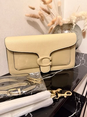 Beige väska från Coach - Stilren beige kuvertväska från Coach i skinn med klassisk C-detalj på locket. Väskan har avtagbar axelrem i matchande färg och guldfärgade metalldetaljer. Insidan har ett fack med dragkedja för småsaker. Perfekt för att lyfta din outfit.