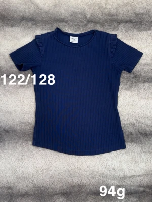 Mörkblå ribbad t-shirt med volang - Snygg mörkblå ribbad t-shirt från Polarn o pyret med volangdetaljer på axlarna. T-shirten har rund halsringning och korta ärmar, perfekt för en trendig vardagslook.🌸 Fint använt skick. Storlek 122/128 