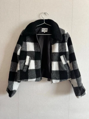 Rutig teddyjacka från NA-KD - Säljer en svartvit rutig teddyjacka från NA-KD med dragkedja framtill och två fickor. Jackan har en bred krage och är gjord i mjukt, fluffigt teddy-material. Perfekt för dig som vill ha en trendig och mysig jacka med coolt rutmönster.