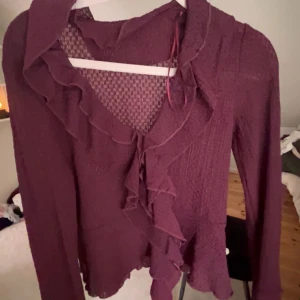 Vinröd volangblus med struktur - Snygg vinröd blus med volangdetaljer längs urringningen och framtill. Blusen har lång ärm och är tillverkad i ett lätt, strukturerat tyg som ger en transparent känsla. Perfekt för dig som gillar romantiska detaljer och en trendig look.