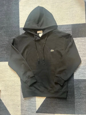 Svart hoodie från Lacoste - Säljer en svart hoodie från Lacoste med klassisk krokodillogo broderad på bröstet. Hoodien har huva med snören, ribbade muddar och en stor magficka. Materialet är mjukt och bekvämt, perfekt för en avslappnad stil.