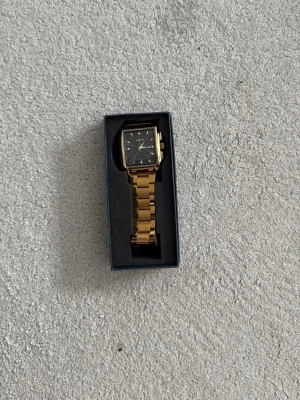 Guldfärgad fyrkantig klocka från LIGE - Stilren guldfärgad klocka från LIGE med fyrkantig svart urtavla och metallarmband. Klockan har tydliga markeringar och en klassisk design som passar till många olika looks. Levereras i originalförpackning.