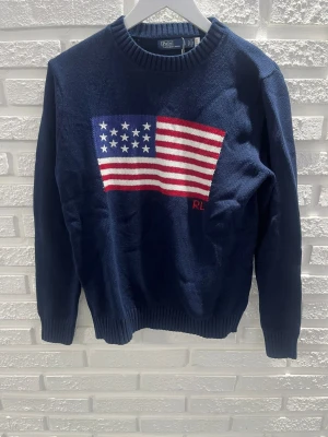 Mörkblå stickad tröja Polo Ralph Lauren - Stickad mörkblå tröja från Polo Ralph Lauren med amerikansk flagga i rött, vitt och blått på bröstet samt RL-broderi. Tröjan har rund halsringning och ribbade muddar vid ärmslut och nederkant. Perfekt för dig som gillar klassisk college-stil.
