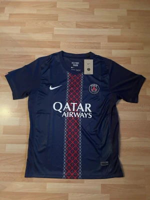 PSG matchtröja Nike blå röd - Snygg PSG matchtröja från Nike i mörkblått med röd och vit grafisk detalj i mitten. Tröjan har klubbmärke och Nike-logga på bröstet samt Qatar Airways som sponsortryck. Tillverkad i lätt och ventilerande Dri-FIT material, perfekt för fotboll eller träning.