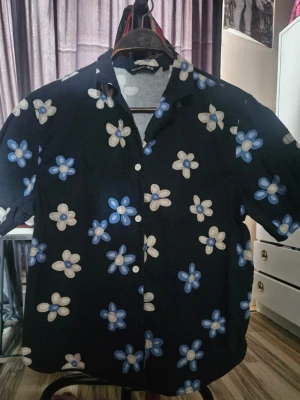 Marimekko skjorta med kort ärm - Snygg mörkblå skjorta med kort ärm och stora blommor i blått och vitt. Storlek M Skjortan har klassisk krage och knappar framtill. Perfekt för dig som vill sticka ut med ett lekfullt mönster. Materialet känns lätt och luftigt, perfekt för varmare dagar.