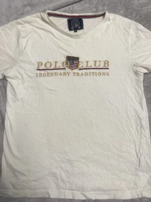 Vit t-shirt från Vinson Polo Club - Vit t-shirt från Vinson Polo Club med broderad logga och texten 'Legendary Traditions' på bröstet. Klassisk rund halsringning och korta ärmar. Perfekt för en avslappnad och stilren look.