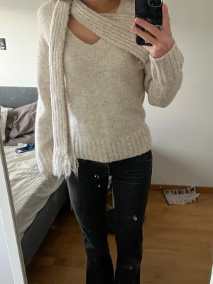 Beige stickad tröja med scarfdetalj -  Trendig beige stickad tröja med unik scarfdetalj över axeln och v-ringning. Tröjan har långa ärmar och ribbstickade muddar vid ärmslut och nederkant.