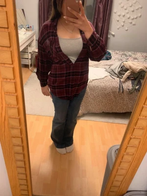 Rutig vinröd skjorta i bomull - Säljer en rutig vinröd skjorta med svarta och vita detaljer. Skjortan har lång ärm, klassisk krage och knappar framtill. Tillverkad i mjuk bomull och har en avslappnad passform som är perfekt för en chill look.