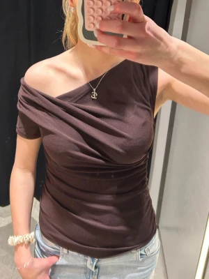 Zara off-shoulder topp - Säljer en brun/mörklila oneshoulder topp med kort ärm och draperad detalj framtill. Toppens material är stretchigt och sitter tight mot kroppen. Perfekt för dig som gillar en trendig och unik look.
