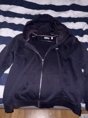 Mörkblå hoodie från GANT med dragkedja - Snygg mörkblå hoodie från GANT med dragkedja framtill och klassisk huva. Tröjan har två fickor och en liten GANT-logga på bröstet. Insidan är mjuk och skön, perfekt för chill dagar. Detaljer med röd och vit rand på insidan av kragen.