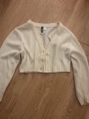Vit cropped kofta från Hm - Säljer en vit cropped kofta från Hm med v-ringning och ribbade detaljer. Koftan har långa ärmar och stängs med fyra stora knappar framtill. Perfekt att styla över ett linne eller t-shirt för en trendig look.