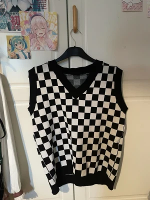 Svartvit rutig stickad väst - Säljer en svartvit stickad väst med schackrutigt mönster och v-ringad hals. Perfekt att styla över en skjorta eller t-shirt för en trendig look. Västen har ribbade kanter vid hals och ärm, och är loose fit för en avslappnad vibe. Den är köpt från Shopee, ca ~150-200kr för flera år sedan. Knappt använd utan defekter.