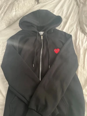 Svart Comme des Garçons Play hoodie - Svart hoodie från Comme des Garçons Play med dragkedja framtill och huva med snörning. På bröstet sitter det ikoniska röda hjärtat med ögon. Hoodien har ribbade muddar och fickor på sidorna. Perfekt för en chill och stilren look.