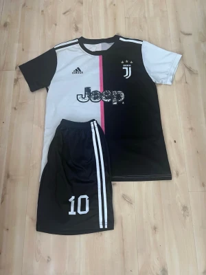 Juventus matchställ svart/vit Adidas - Juventus matchställ med svart och vit t-shirt och shorts. Tröjan har rosa rand i mitten, Juventus-logga, Adidas-logga och Jeep-tryck. Shortsen är svarta med vita ränder och nummer 10 tryckt på benet. Perfekt för fotbollsträning eller match.