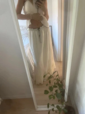 Vit långkjol med volang och snörning - Långkjol med volangkant och svart snörning i midjan i off-white färg. Kjolen har en luftig och flowy passform, perfekt för sommaren. Materialet känns lätt och svalt, och kjolen har en feminin look med fina detaljer. Nypris 749 kr