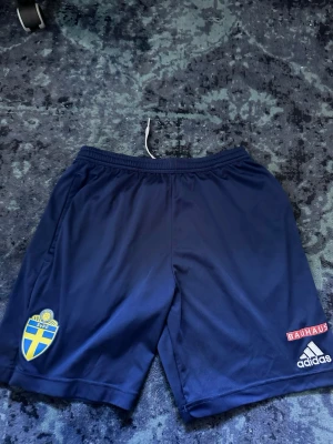 Blå Sverige fotbollsshorts Adidas - Mörkblå fotbollsshorts från Adidas med Svenska Fotbollförbundets märke och Bauhaus-logga. Shortsen har resår i midjan och är tillverkade i ett lätt, snabbtorkande material som passar perfekt till träning eller match.