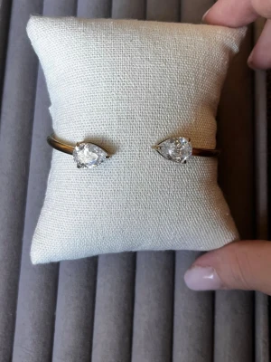 Guldfärgat armband med droppformade stenar - Snyggt guldfärgat armband i metall med två stora, droppformade klara stenar i varje ände. Öppen design som gör det lätt att ta på och av. Perfekt för dig som gillar stilrena och eleganta accessoarer med lite extra bling.