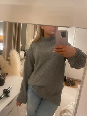 Grå oversized stickad tröja - Mysig grå stickad tröja med oversized passform och ribbade muddar vid ärmslut och nederkant. Tröjan har en högre rund halsringning och är perfekt för lager på lager. Passar till jeans eller kjol för en avslappnad look.