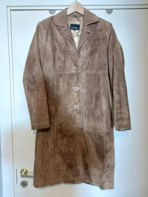 Brun mockakappa från Josefssons - Snygg brun mockakappa från Josefssons med klassisk krage och knäppning framtill. Kappan har en rak passform och är tillverkad i mjukt mockamaterial som ger en cool vintagekänsla. Perfekt för dig som vill ha en tidlös och stilren look.
