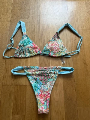 Bikini - Säljer en superfin bikini med paisleymönster i flera färger som rosa, blått, grönt och gult. Både överdel och underdel har ljusblå band och knyt. Materialet är mjukt och stretchigt i polyester och elastan. Perfekt för stranden eller poolen!