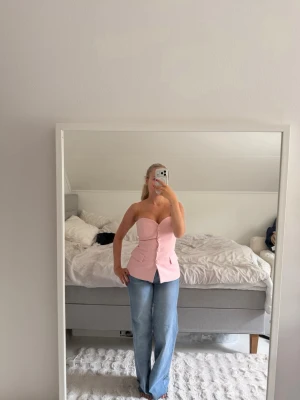 Ljusrosa axelbandslös korsettopp - Trendig ljusrosa korsettopp med markerad midja och knappar framtill. Axelbandslös design som ger en snygg siluett och passar perfekt till jeans eller kjol. Aldrig använt! Kostade 500kr 