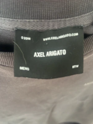 Svart t-shirt från Axel Arigato - Cool svart t-shirt från Axel Arigato med tryck på ryggen som visar 'ARIGATO ©2014' och adressdetaljer. Klassisk passform och rund halsringning. Perfekt för dig som gillar stilrena och moderna plagg med en twist.