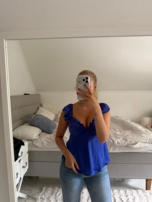 Blå v-ringad topp med volangärm - Snygg blå topp med v-ringning och volangdetaljer vid ärmarna. Toppen har en luftig passform och är tillverkad i ett lätt material som ger ett avslappnat intryck. Perfekt till jeans eller kjol för en trendig look.