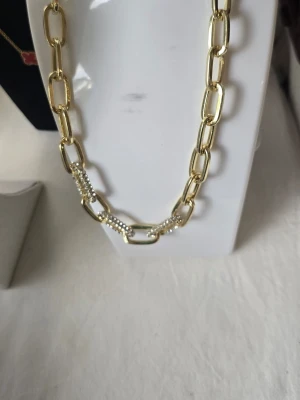 Guldigt halsband med pärldetaljer - Snyggt halsband i guldfärgad metall med stora, rektangulära länkar. Några av länkarna är dekorerade med små vita pärlor som ger en extra touch. Perfekt för dig som vill ha ett statement-smycke med trendig känsla.