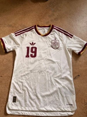 Spanien Lamine Yamal matchtröja - Vit Spanien matchtröja från Adidas med vinröda detaljer, nummer 19 och Lamine Yamal på ryggen. Tröjan har klassiska Adidas-ränder på axlarna, broderad landslagssköld och gul kant vid hals och ärmslut. Tillverkad i lätt funktionsmaterial, perfekt för fotboll.