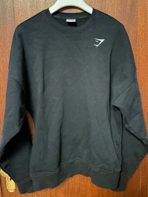 Svart oversized sweatshirt från Gymshark - En stilren svart sweatshirt från Gymshark med rund halsringning och diskret logga på bröstet. Tröjan har långa ärmar och ribbade muddar vid ärmslut och nederkant. Perfekt för en avslappnad och sportig look.