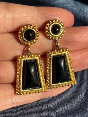 Vintage Monet Svart Guldiga Örhängen - Unika örhängen i svart och guld med geometrisk design. Överdelarna är runda med svarta stenar, och underdelarna är trapetsformade med svarta infattningar. Guldfärgad metall med detaljerade kanter ger en lyxig retrovibe. Perfekta för dig som gillar statement-smycken. I fint skick. 