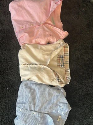 3 skjortor från Morris Lady & Burberry - Säljer tre skjortor: en ljusrosa Morris Lady med klassisk krage och knappar, en vit Burberry med rutigt tyg på insidan av knappslån och en ljusblå skjorta med diskreta ränder. Alla har lång ärm och tidlös design. Perfekta till en clean och fräsch stil.