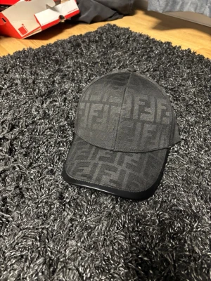 Svart keps från Fendi med logomönster - Snygg svart keps från Fendi med det klassiska FF-logomönstret över hela kepsen. Kepsen har böjd skärm och är tillverkad i ett tygmaterial med en stilren look. Perfekt accessoar för att lyfta din outfit.