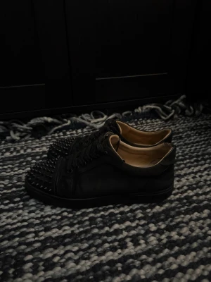 Svarta Louboutin sneakers med nitar - Säljer ett par svarta sneakers från Christian Louboutin med coola nitar på tån och klassisk röd sula. Skorna är låga och har en stilren design i läder med svarta skosnören. Perfekta för dig som vill sticka ut med en edgy look. De är i lite sämre skick tyvärr ingen låda har för mig de är riktiga inte säker dock