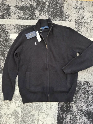 Svart ziptröja från Polo Ralph Lauren - Klassisk svart ziptröja från Polo Ralph Lauren med hög krage och vit broderad logga på bröstet. Tröjan har två fickor framtill och ribbade muddar vid ärmslut och nederkant. Perfekt för en stilren och avslappnad look. OBS: LITEN I STORLEKEN
