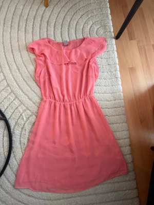 Rosa kortklänning med volang - Söt rosa kortklänning med volangdetalj vid halsen och resår i midjan. Klänningen är ärmlös och har en luftig, flowy passform som passar perfekt till varma dagar. Materialet känns lätt och skönt mot huden.